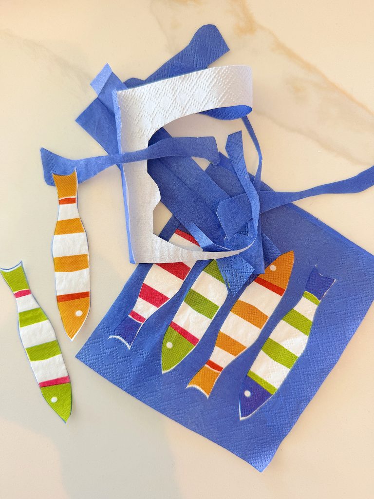 Blue napkin with colorful fish shapes cut out, placed on a white surface; cut-out pieces and fish shapes are arranged nearby.
