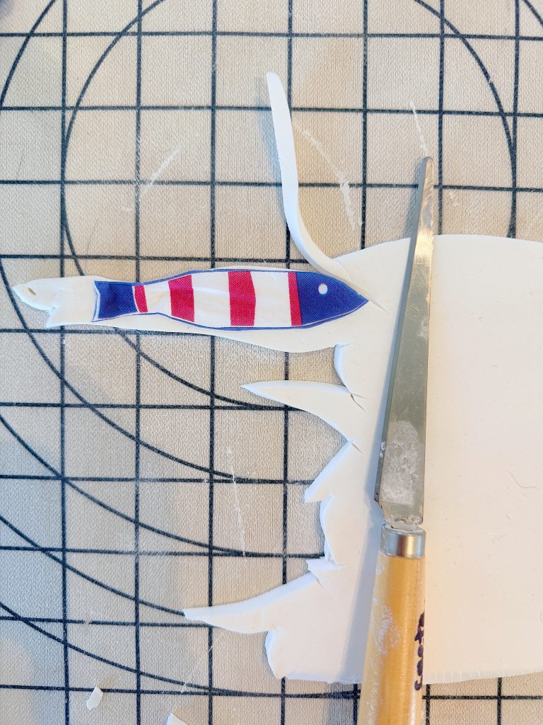A craft knife cuts white material near a small fish-shaped piece with red, white, and blue stripes, placed on a gridded cutting mat.