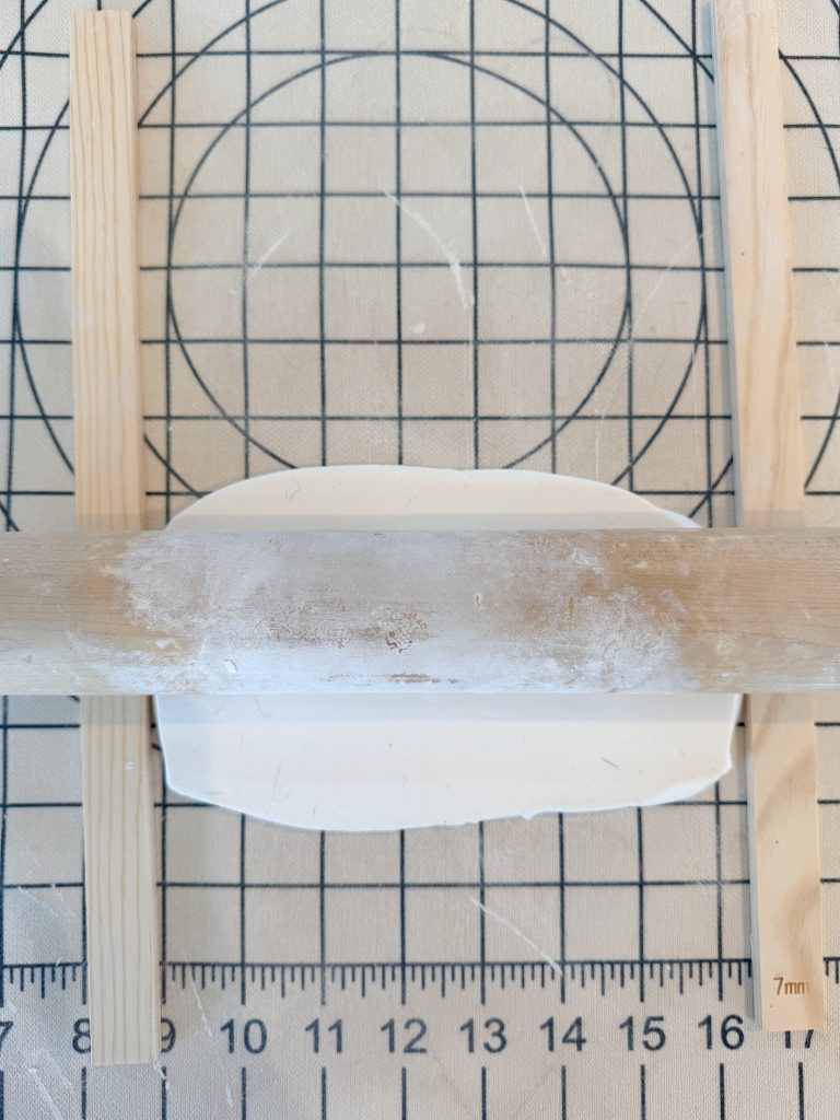 A rolling pin flattens white clay between two wooden guides on a gridded work surface with measurement markings.