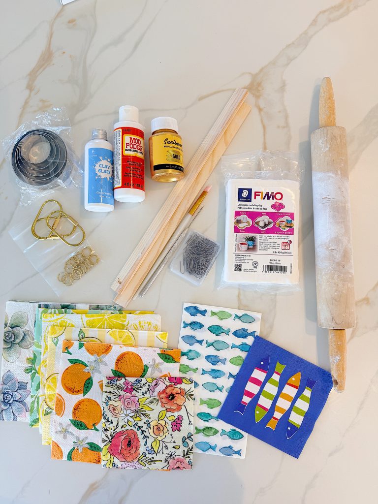 Assorted crafting supplies on a white surface, including napkins, paints, Mod Podge, FIMO clay, wooden sticks, cookie cutters, sandpaper, binder clips, and a rolling pin.