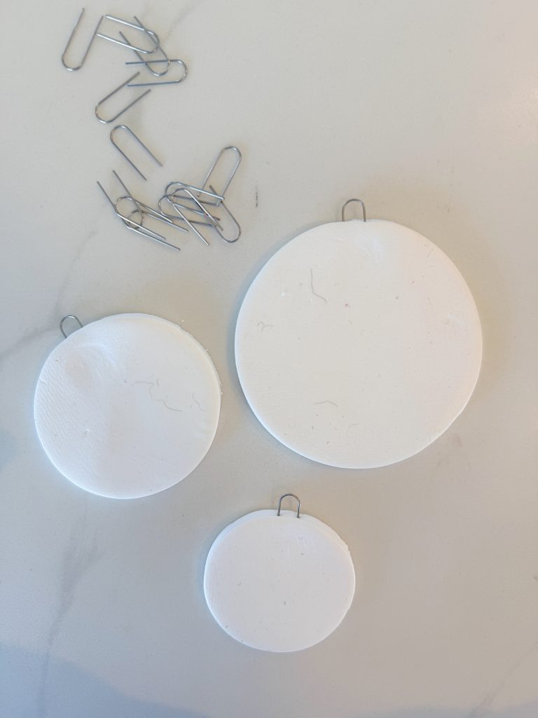 Three round white clay ornaments with paper clips inserted at the top are on a white surface, next to several unused paper clips.