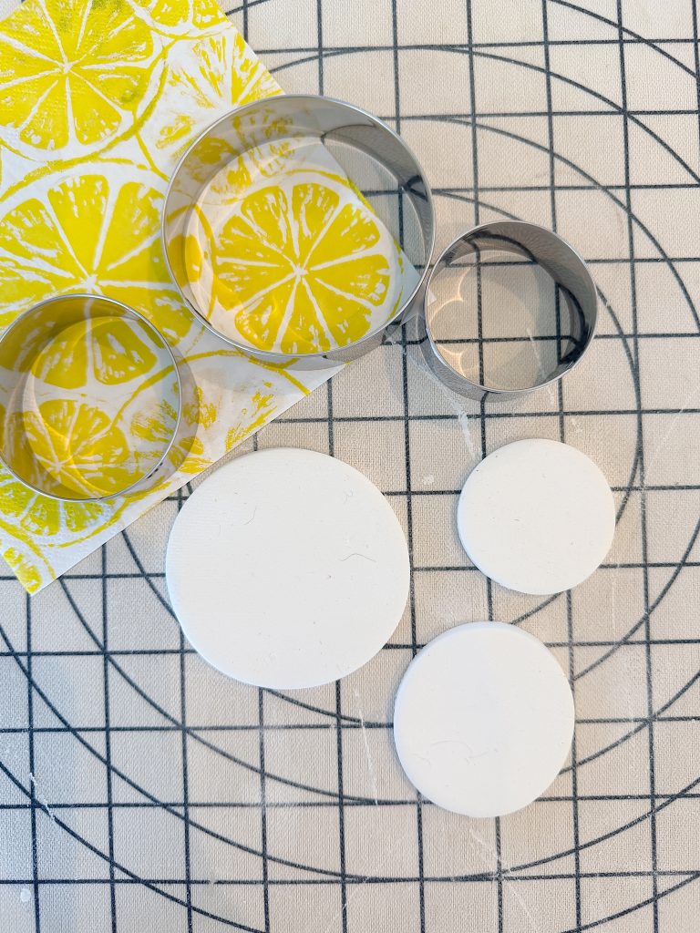 Three metal round cookie cutters, yellow citrus-printed paper, and three white clay circles are on a black grid-patterned surface.