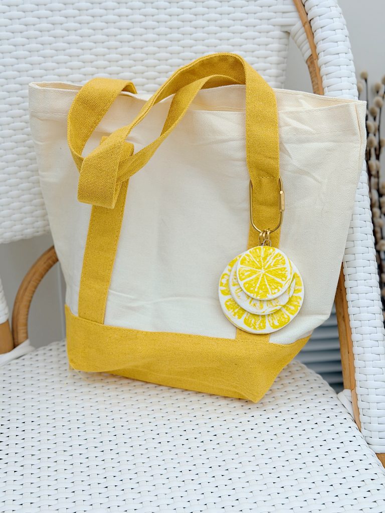 A cream and yellow canvas tote bag with a citrus slice keychain is placed on a white woven chair.