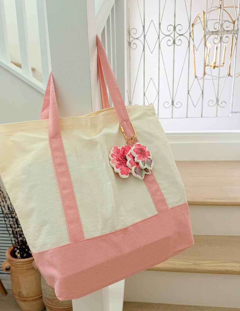 A cream and pink tote bag with pink flower keychains hangs on a white stair railing indoors near wooden steps.