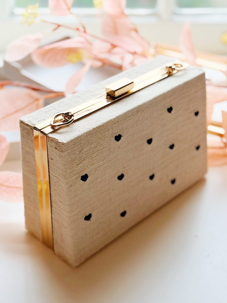 A rectangular beige Box Frame Purse with small black polka dots and gold metal accents, placed on a white surface with pink decorative leaves in the background.