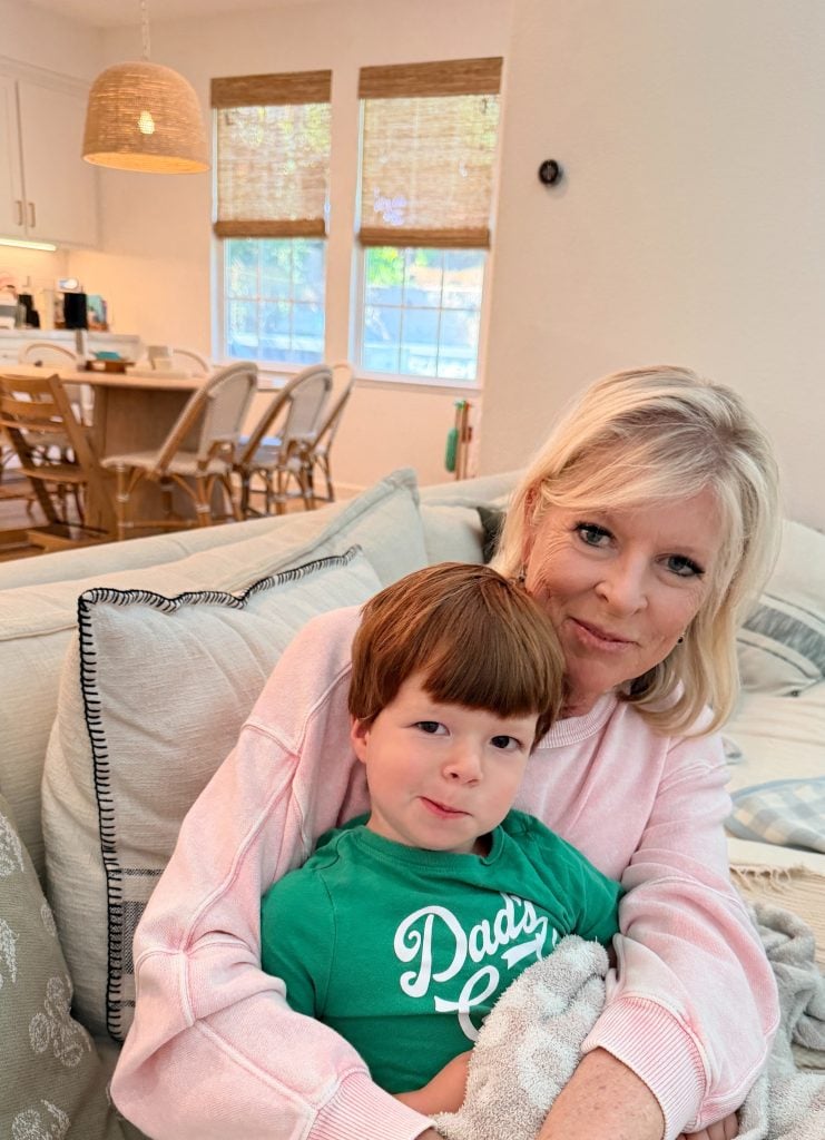 A woman with blonde hair hugs a young boy in a green "Daddy's Golf" shirt while sitting on a couch in a bright, cozy living room with large windows.