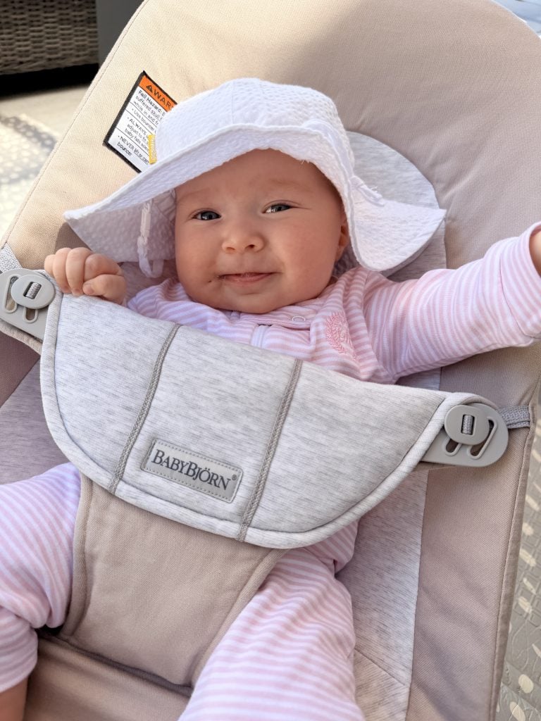Baby in a pink and white striped outfit and white sun hat sits in a beige BABYBJÖRN infant seat, smiling with one hand touching the hat.