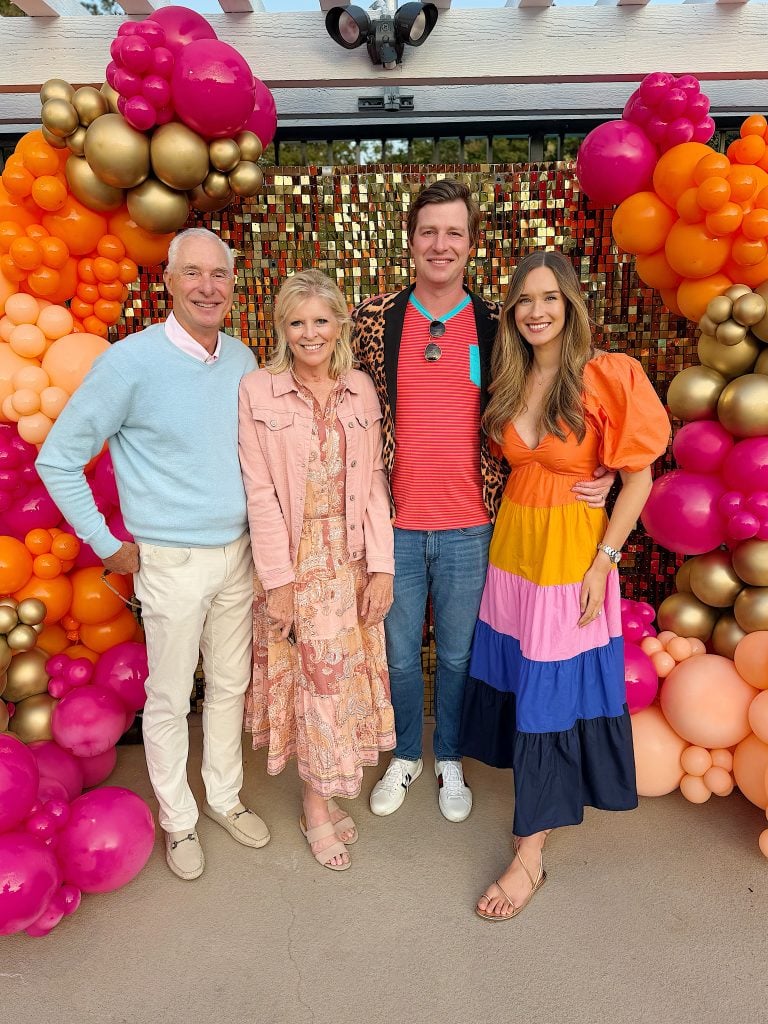 Four people stand smiling in front of a colorful balloon and sequin backdrop, dressed in bright, festive clothing.
