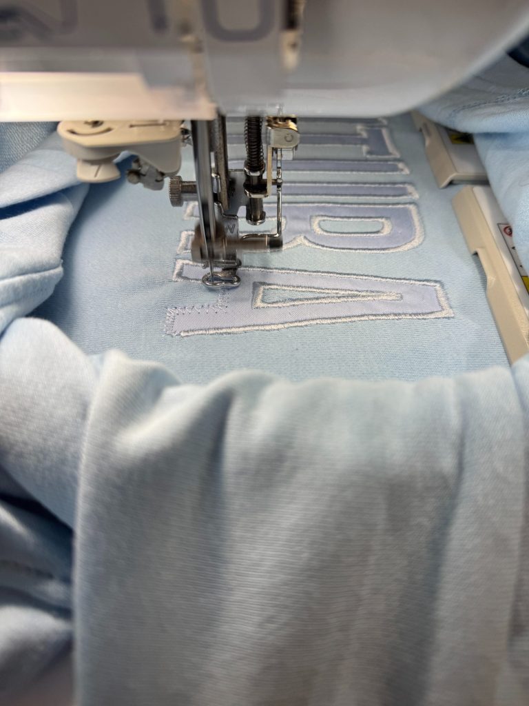 Close-up of an embroidery machine stitching large, block letters onto light blue fabric.