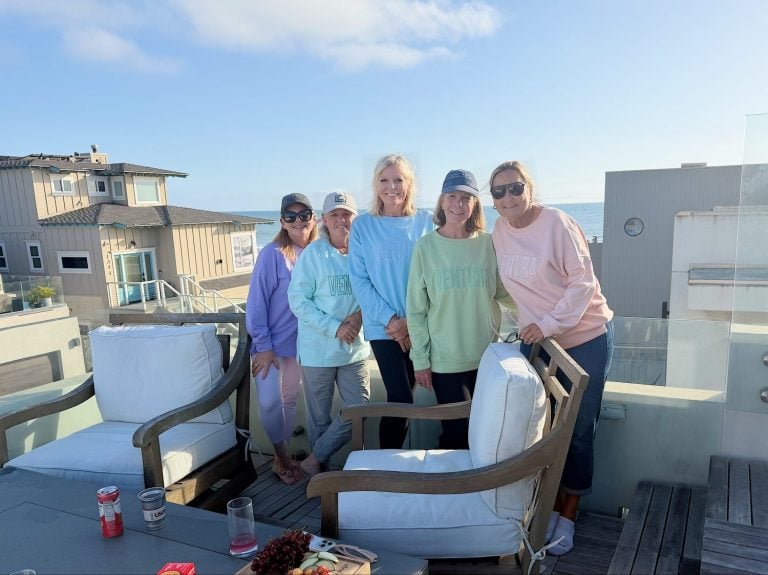 Five women stand and smile on a rooftop patio overlooking the ocean, wearing pastel sweatshirts and hats. Outdoor furniture and snacks are visible in the foreground.