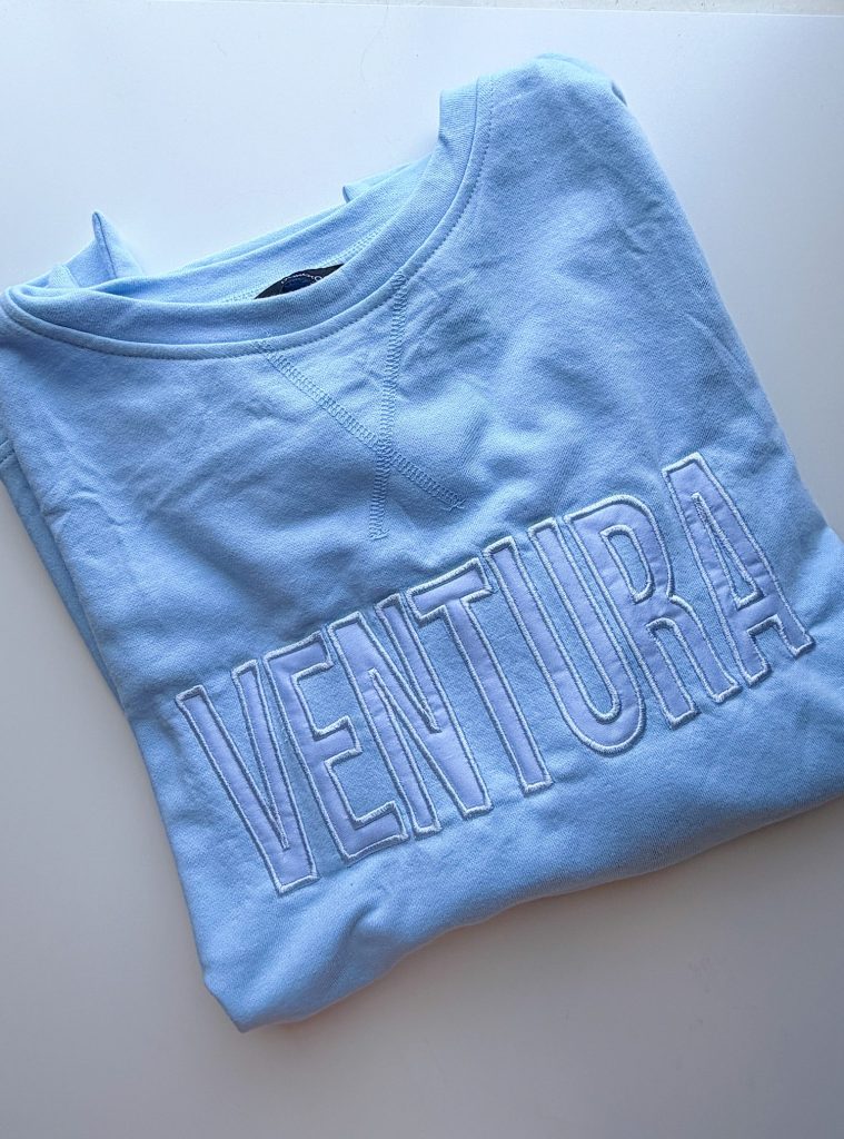 A folded light blue sweatshirt with "VENTURA" embroidered in large capital letters on the front.