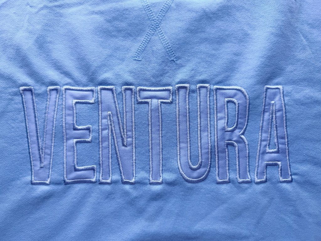 Light blue fabric with the word "VENTURA" embroidered in large outlined capital letters and an "X" stitched above it.