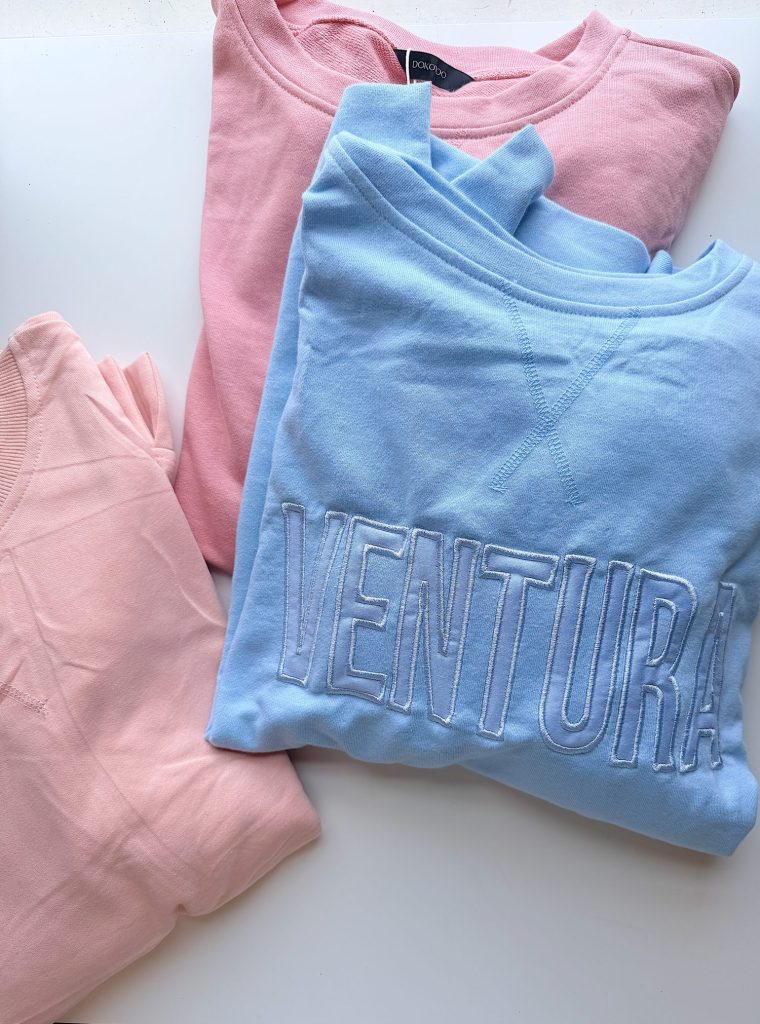 Three folded sweatshirts are shown: two light pink ones and one light blue one with "VENTURA" embroidered in large white letters.