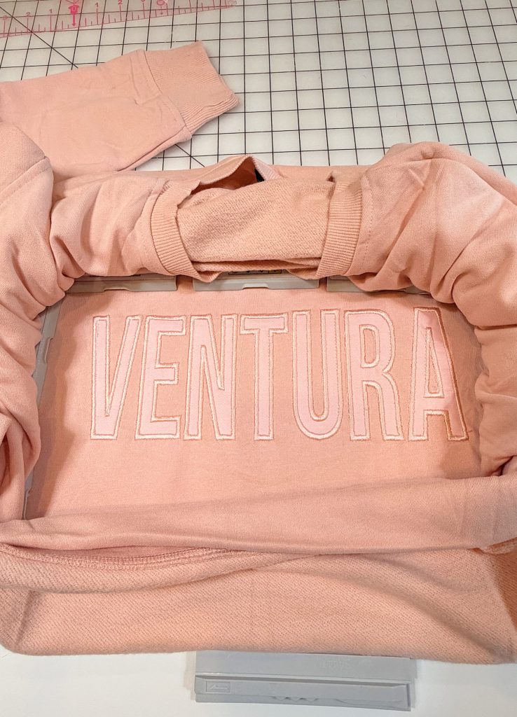 A pink sweatshirt with the word "VENTURA" stitched in large white letters across the front, laid flat on a grid-patterned surface.