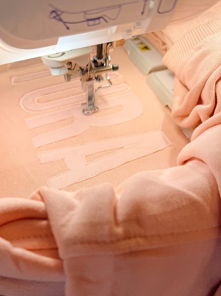 A close-up of a sewing machine embroidering large letters onto a peach-colored fabric or garment.