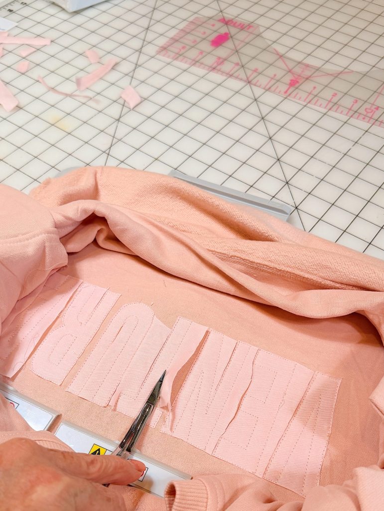 A person cuts vertical slits into fabric letters on a pink sweatshirt with scissors, on a grid-patterned cutting mat.
