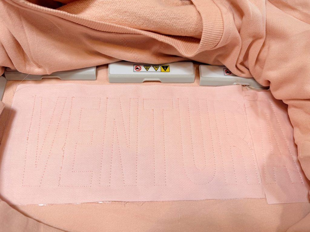 A light pink fabric with the word "VENTURA" outlined in dotted lines, placed under a machine with caution labels.
