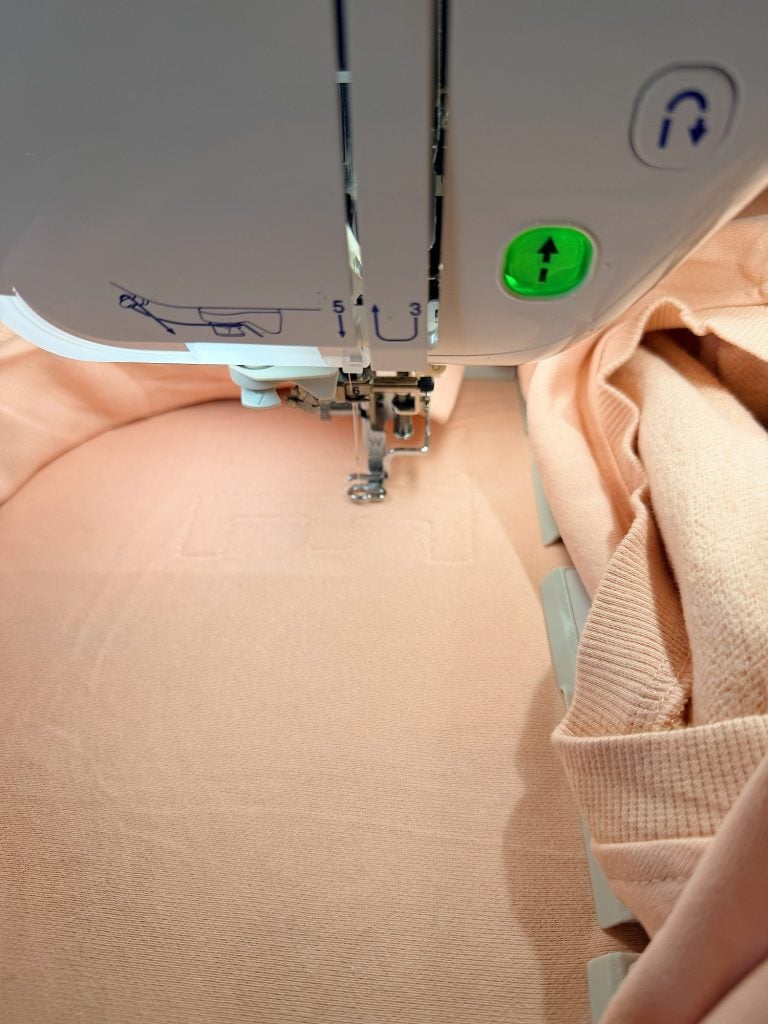 Close-up of a sewing machine stitching a rectangular pattern onto peach-colored fabric. A green light indicates the machine is operating.