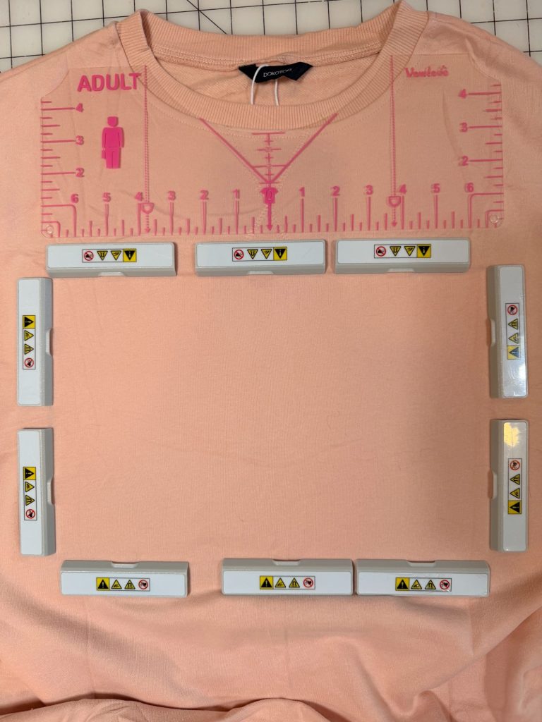 A peach-colored shirt with a pink positioning ruler and a rectangular alignment frame with warning labels placed on the chest area for precise design placement.