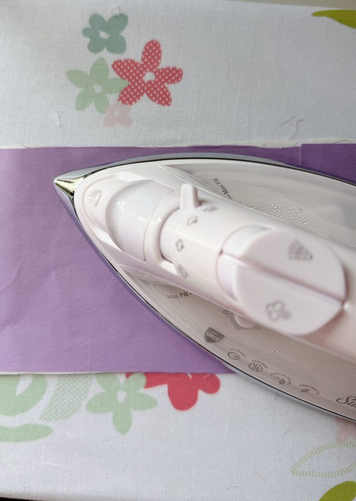 A white iron presses a strip of purple fabric on a floral-patterned ironing board cover.
