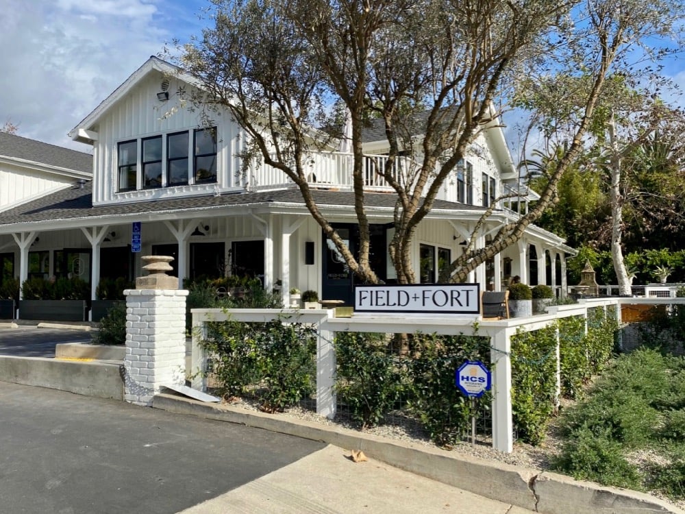 A white, two-story building with a porch and trees in front, features a sign reading "FIELD + FORT" and is bordered by a white fence and greenery.