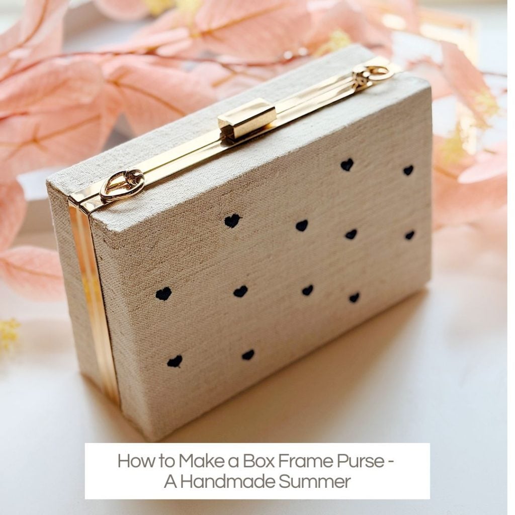 A rectangular box frame purse with gold hardware and a light fabric exterior featuring small black dots, placed near pink decorative leaves.