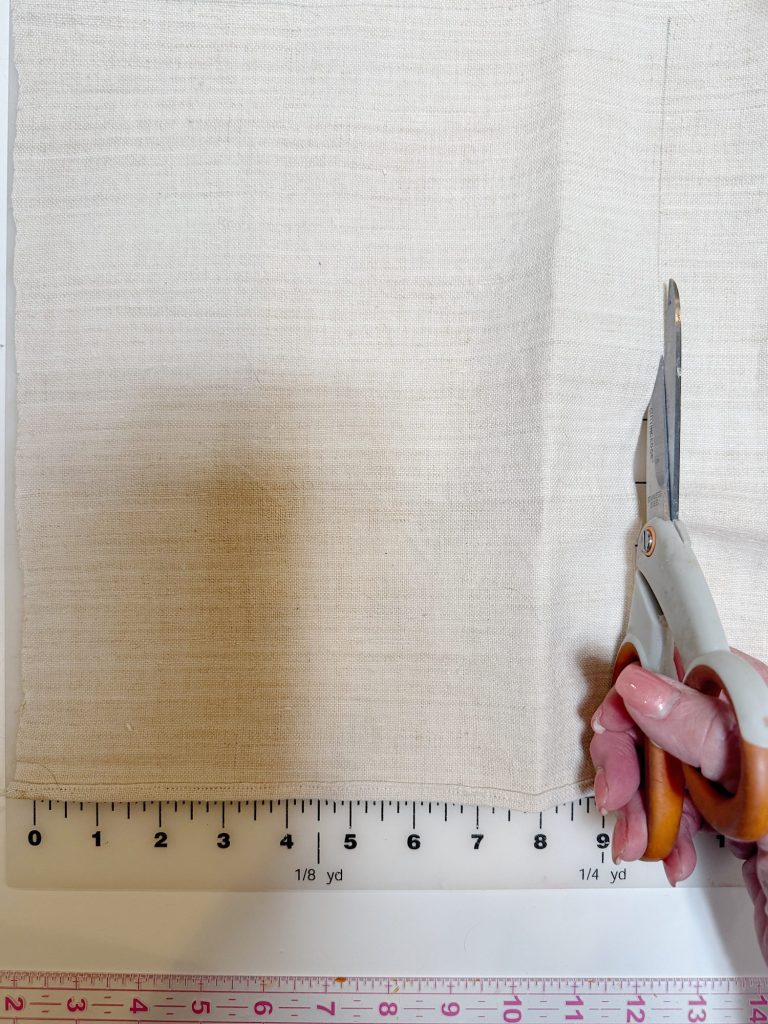 A person cuts a piece of off-white fabric with scissors along a line, beside a ruler showing measurements in inches and yards.