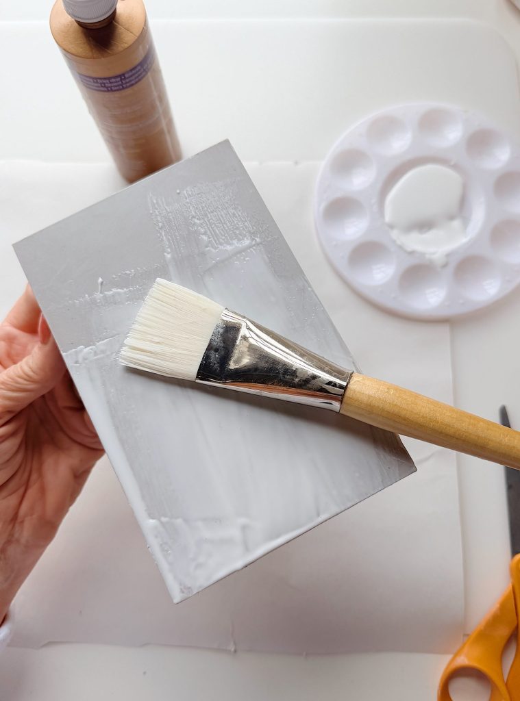 A hand holds a rectangular object while a paintbrush applies white paint to its surface; nearby are a paint bottle, a palette with white paint, and a pair of scissors.
