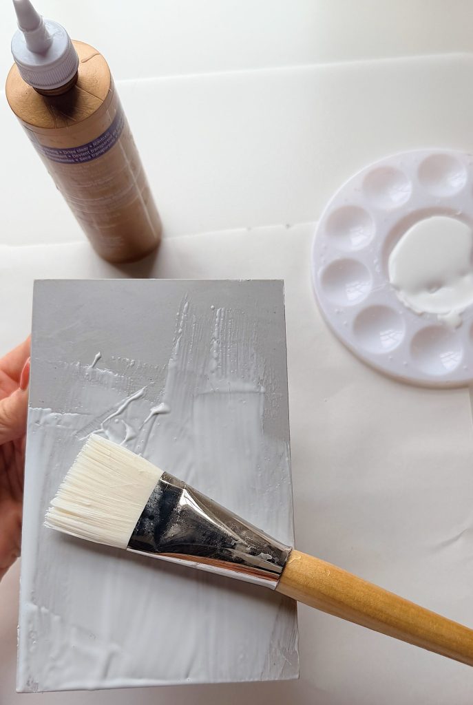 A hand holds a grey rectangular board being coated with white glue using a wide paintbrush, with a glue bottle and a paint palette nearby.