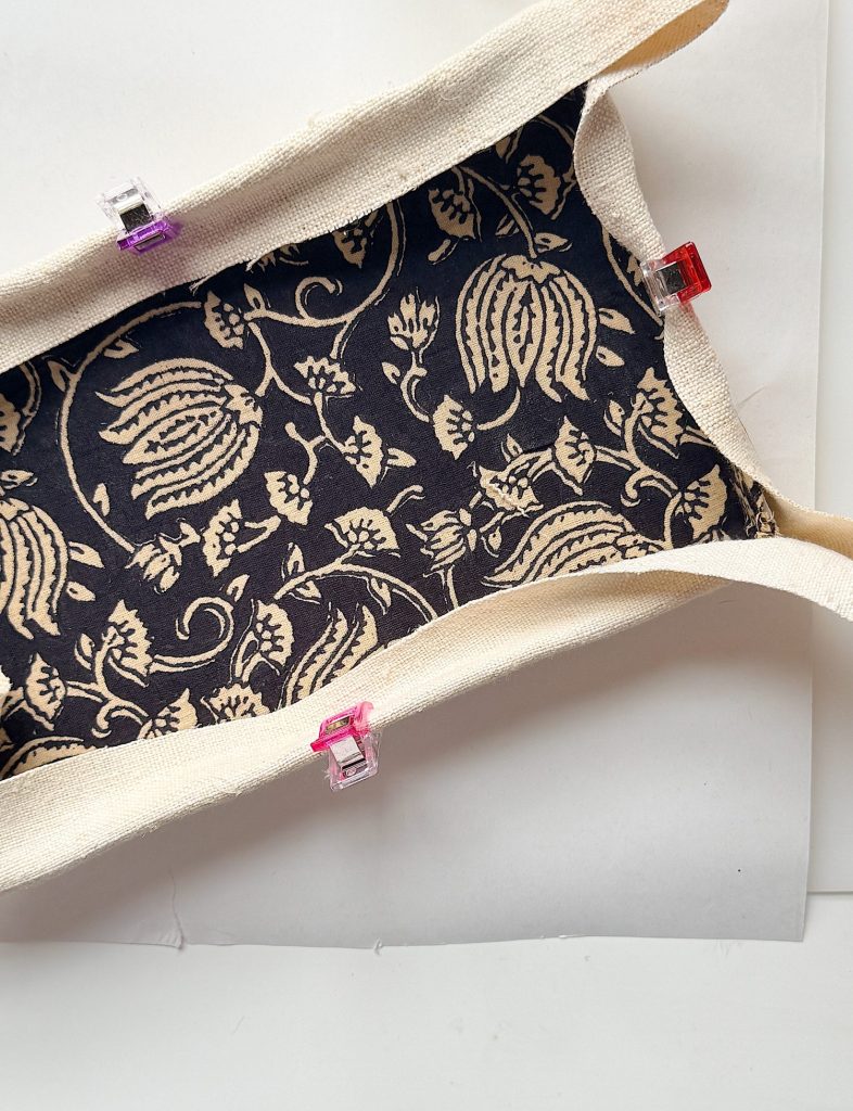 A fabric pouch with a dark floral interior and light outer fabric is held open with three colored clips on the edges, placed on a white surface.