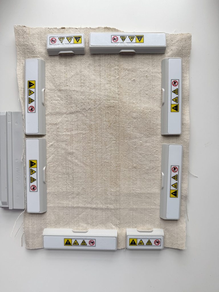 Seven white plastic objects with various yellow and red warning labels are arranged in a rectangular shape on a piece of beige fabric on a white surface.