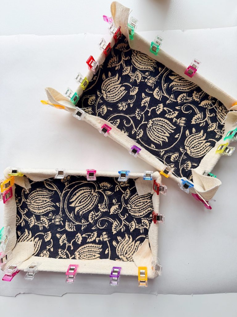 Two rectangular fabric trays with patterned navy and beige fabric are held together at the edges with colorful sewing clips, displayed on a white surface.