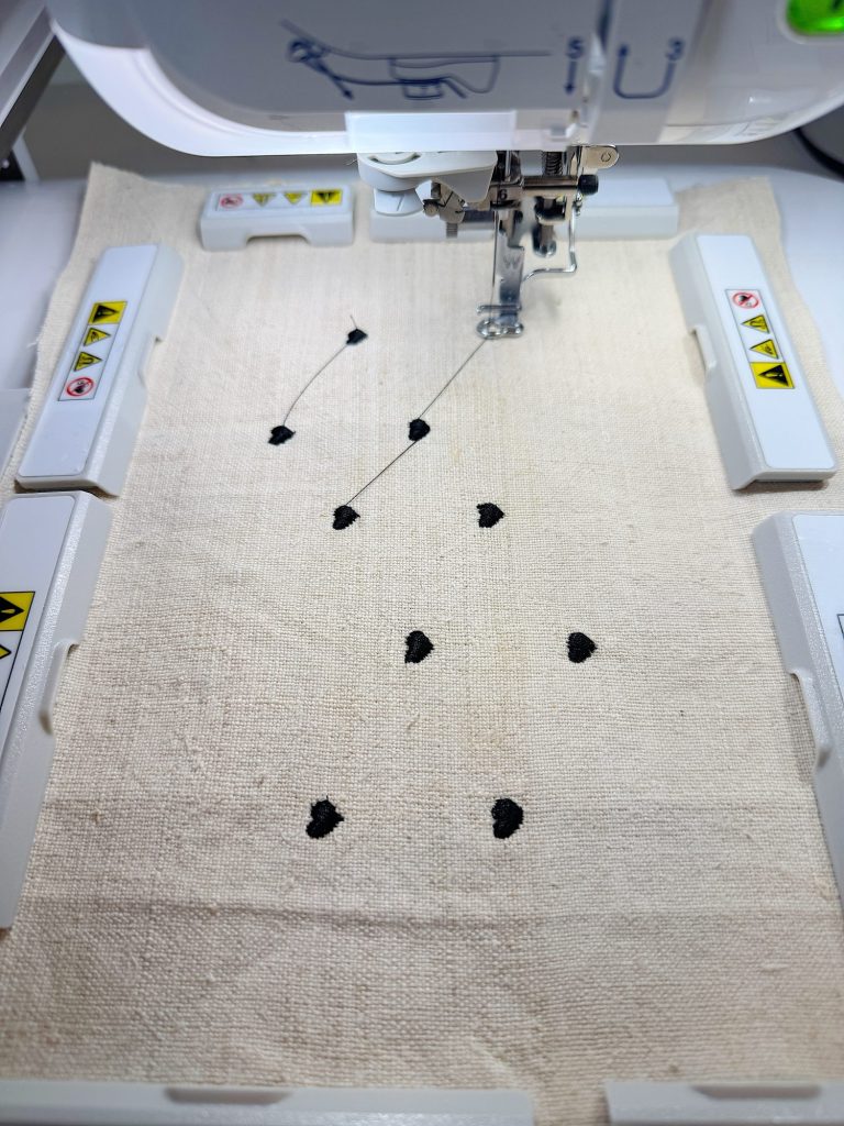Close-up of an embroidery machine stitching small black shapes onto a piece of off-white fabric held in a rectangular frame.