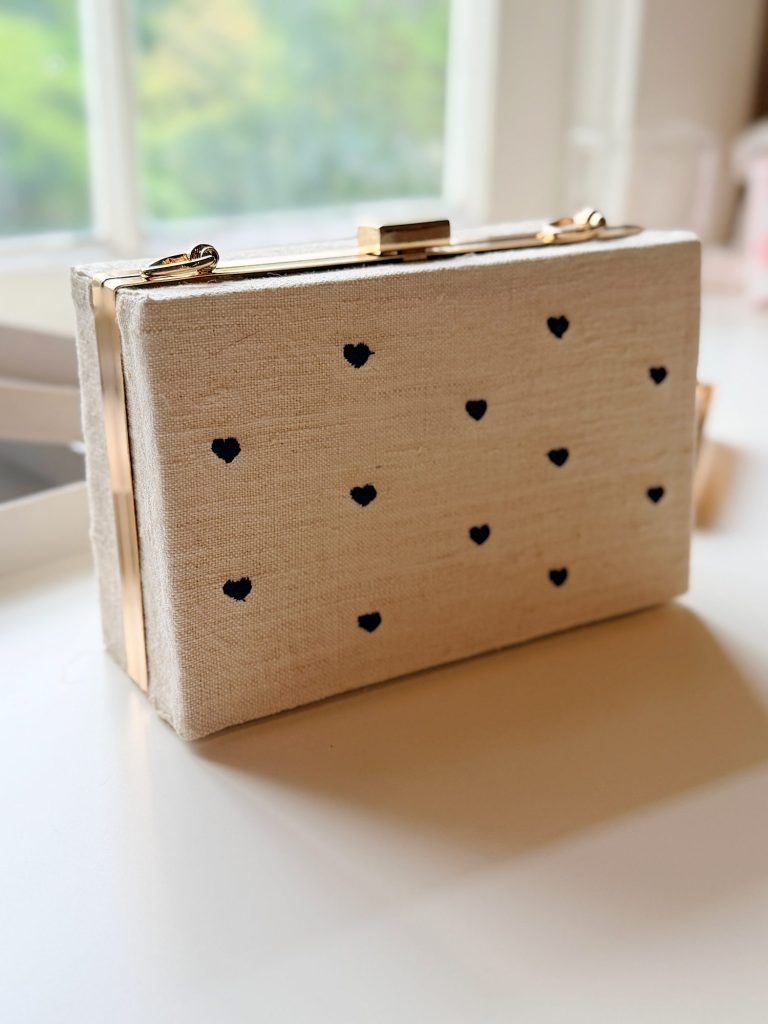 A rectangular beige clutch with a gold clasp and small black heart patterns sits on a white surface near a window.