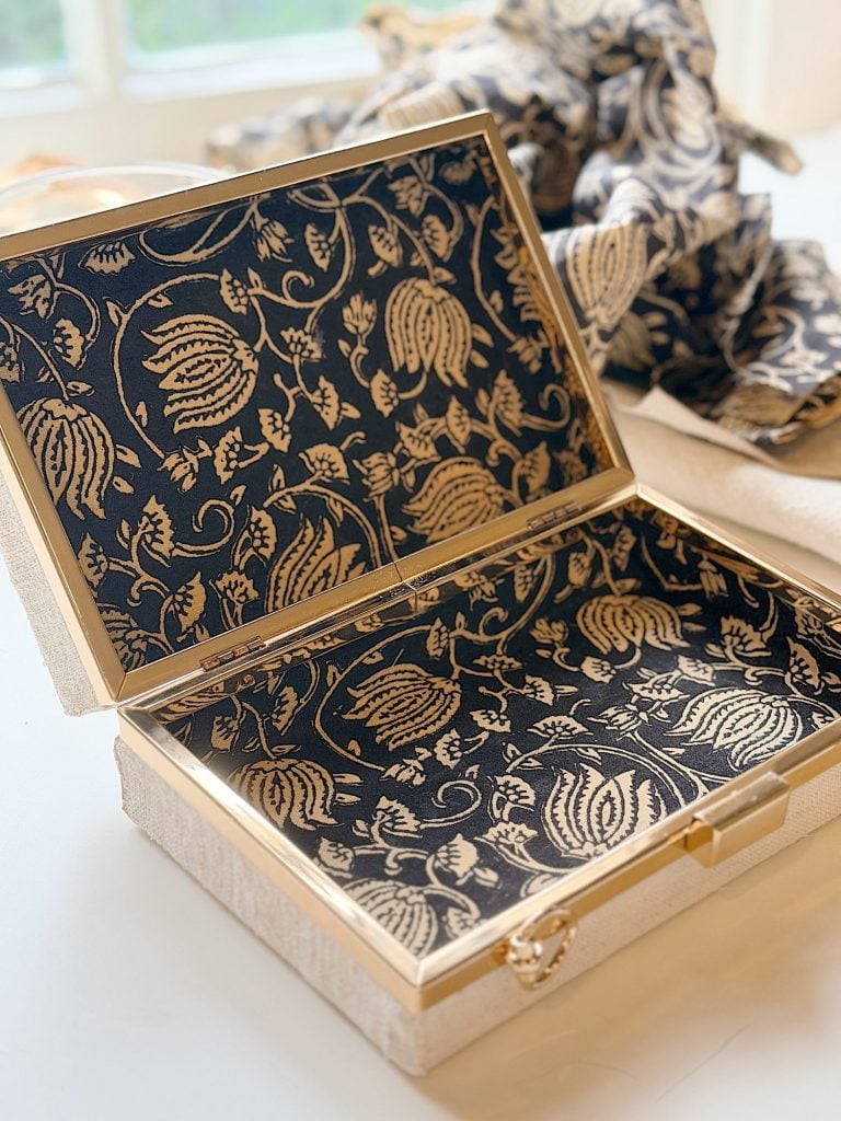 An open rectangular box with gold trim and a black and gold floral patterned interior, placed on a white surface with fabric in the background.