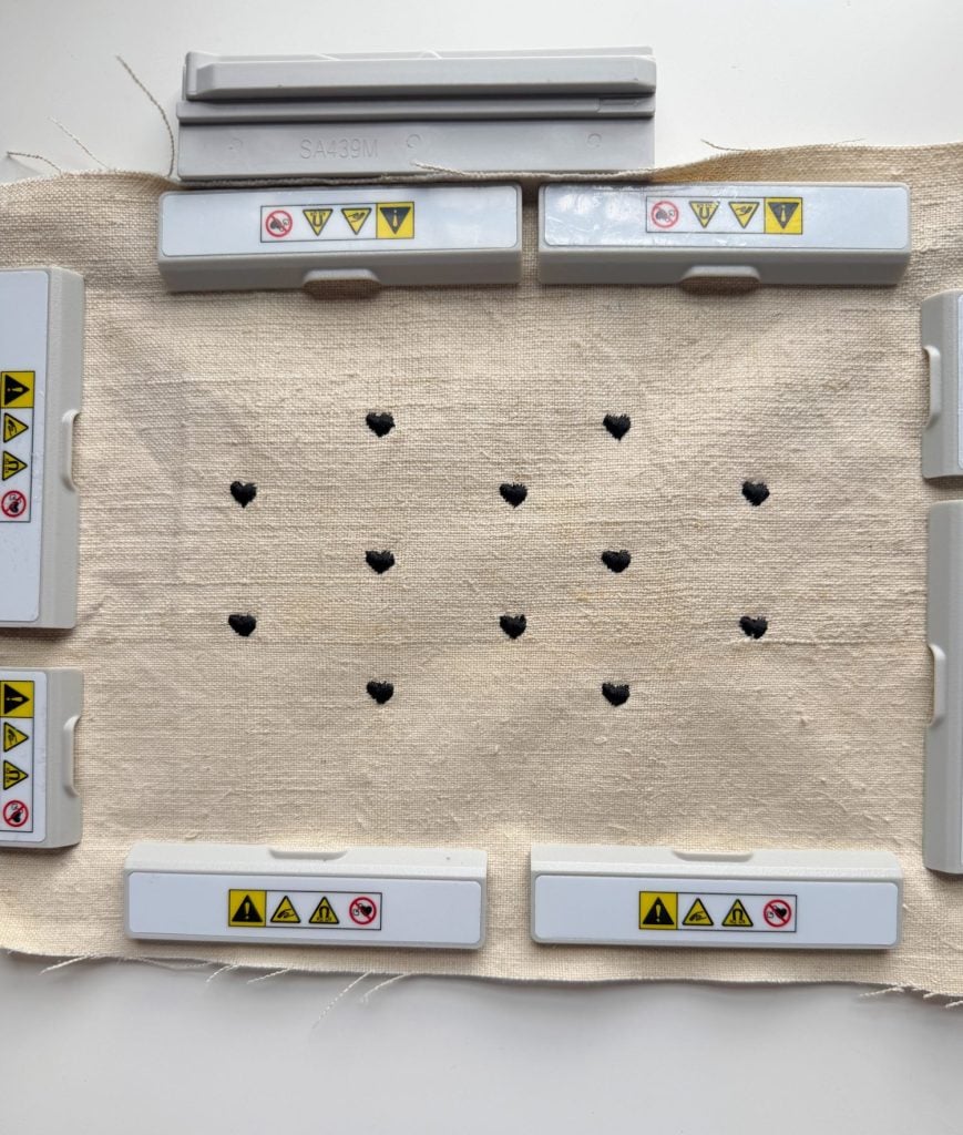A rectangular piece of beige fabric with black heart-shaped cutouts is held in place by gray plastic clamps labeled with caution symbols.