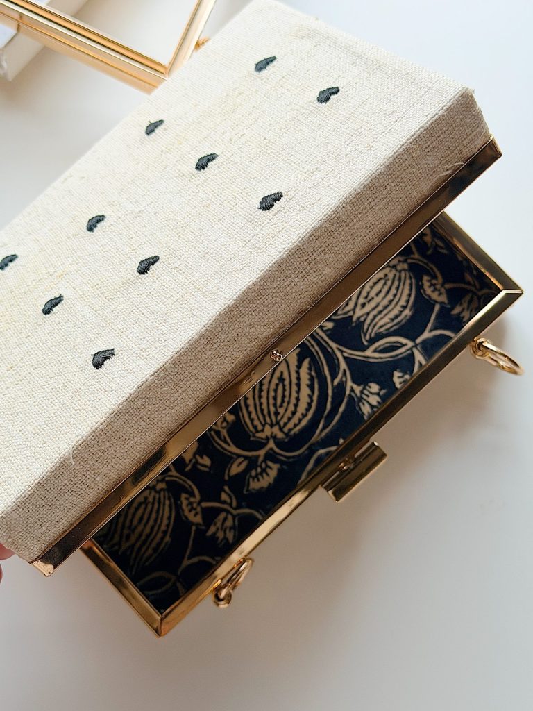 A rectangular clutch with a beige fabric lid featuring small black dots, and a gold frame with a floral-patterned interior lining.