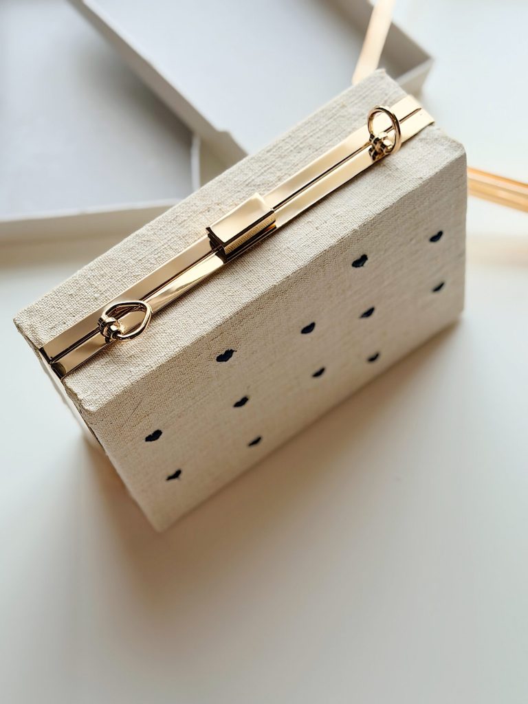 Rectangular beige clutch with small black polka dots, gold metal clasp, and a thin strap, placed on a white surface near an open box.