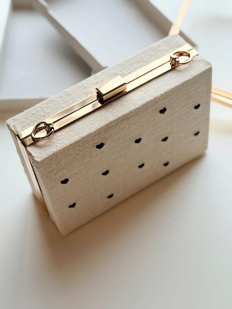 Beige rectangular clutch with a gold metal clasp and small black dots, resting on a white surface near an open box.