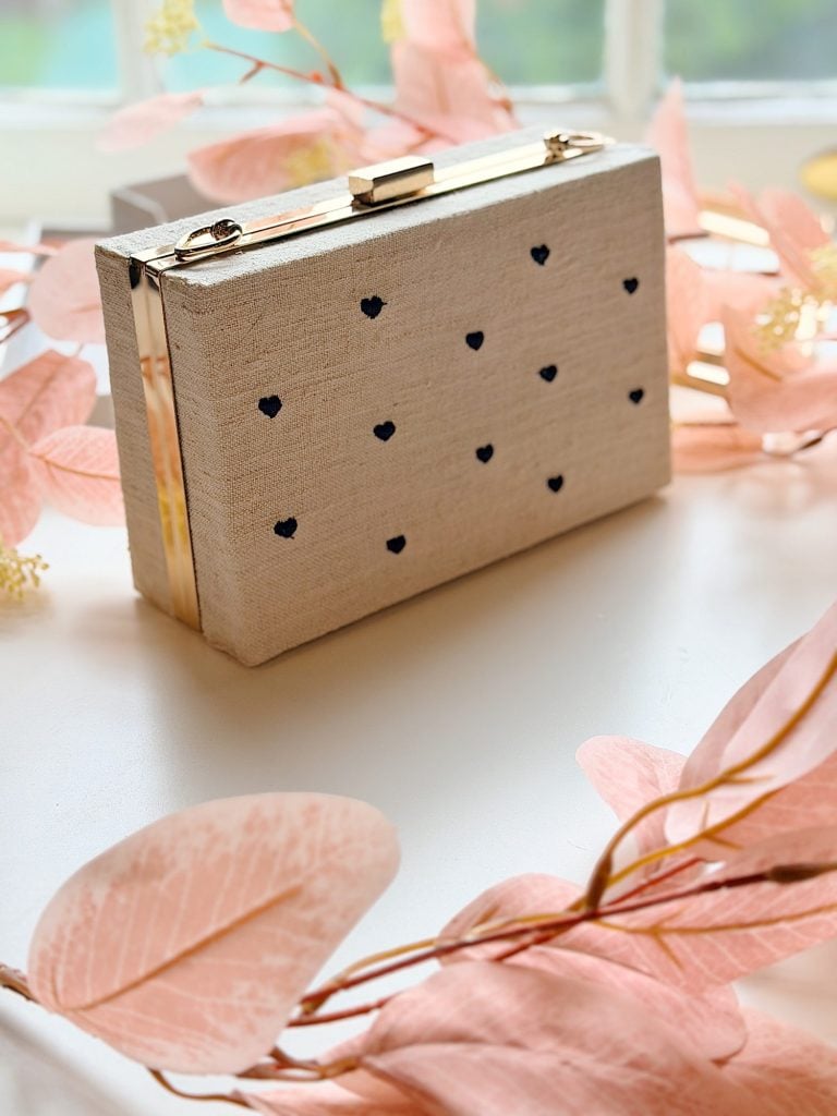 A beige rectangular clutch with small black heart patterns and gold accents is displayed among pink decorative leaves on a white surface.