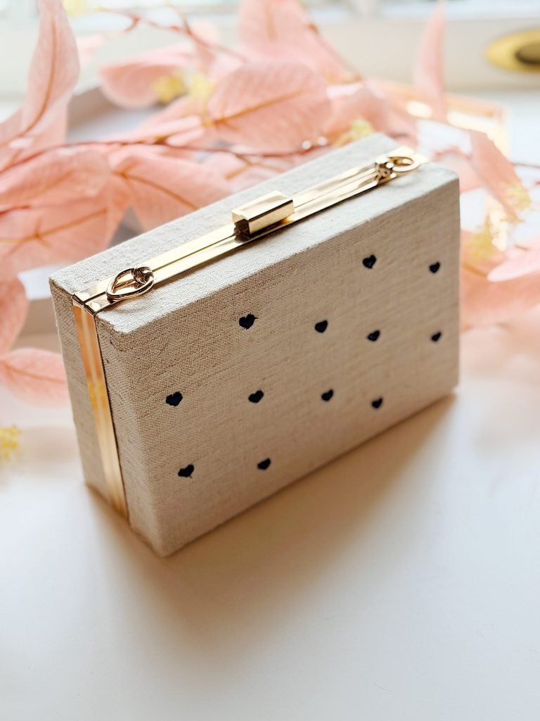 A beige rectangular clutch purse with gold metal accents and small black heart patterns, placed on a white surface with pink decorative leaves in the background.