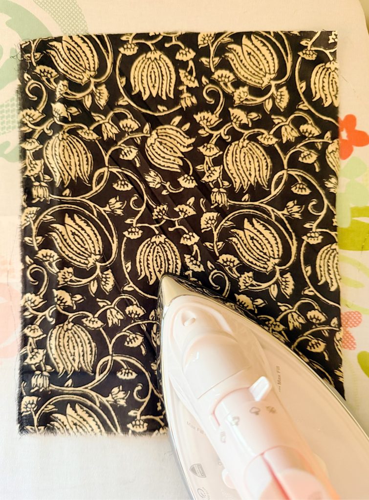 An iron is pressed onto a rectangular piece of black fabric with a white floral pattern, on an ironing board.