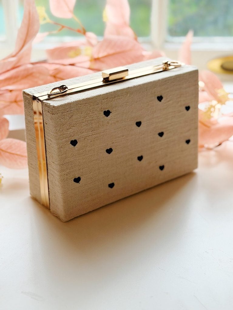 A rectangular beige clutch with a gold clasp and small black heart patterns, placed on a white surface near light pink leaves.
