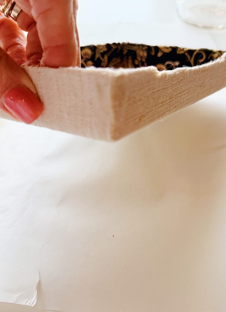 A hand with pink nail polish holds the corner of a rectangular, fabric-covered object with visible stitching and a patterned interior.