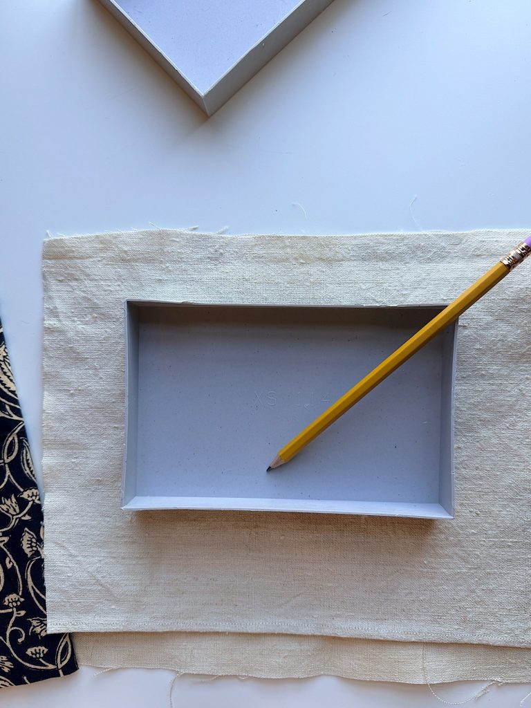 A rectangular gray tray is placed on a piece of off-white fabric, with a yellow pencil resting on the tray&rsquo;s edge. A patterned blue cloth is partially visible on the left.