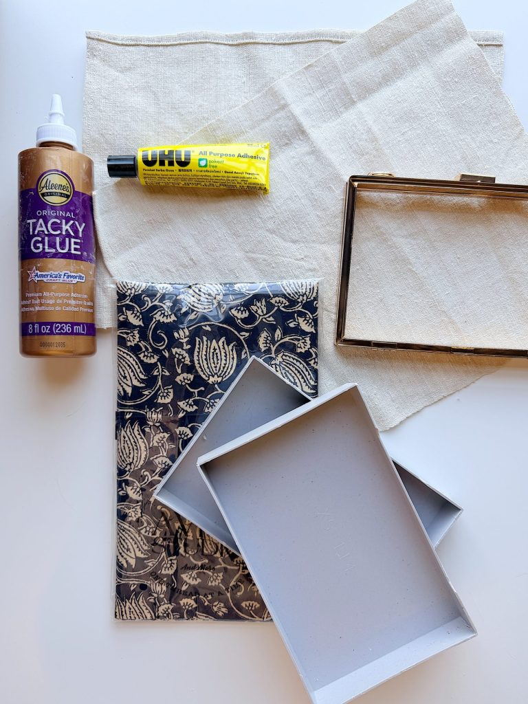 Materials for a craft project, including a bottle of tacky glue, a tube of all-purpose adhesive, fabric pieces, a glass frame, decorative paper, and two gray box lids.