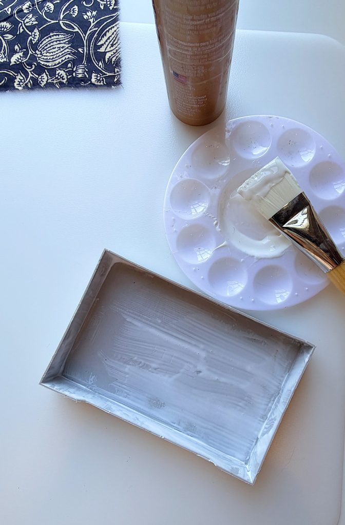 A rectangular tray coated with white glue beside a round paint palette with glue, a brush, and a brown bottle on a white surface.