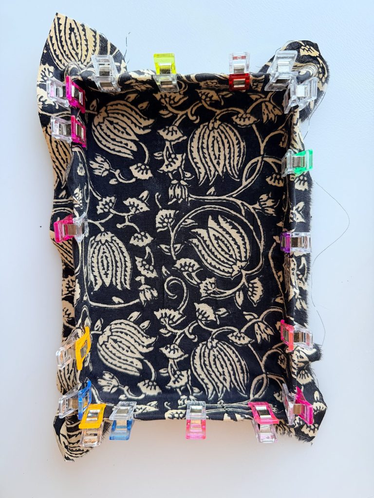A rectangular fabric piece with a black and beige floral pattern is clipped at the edges with colorful plastic sewing clips, forming raised sides.