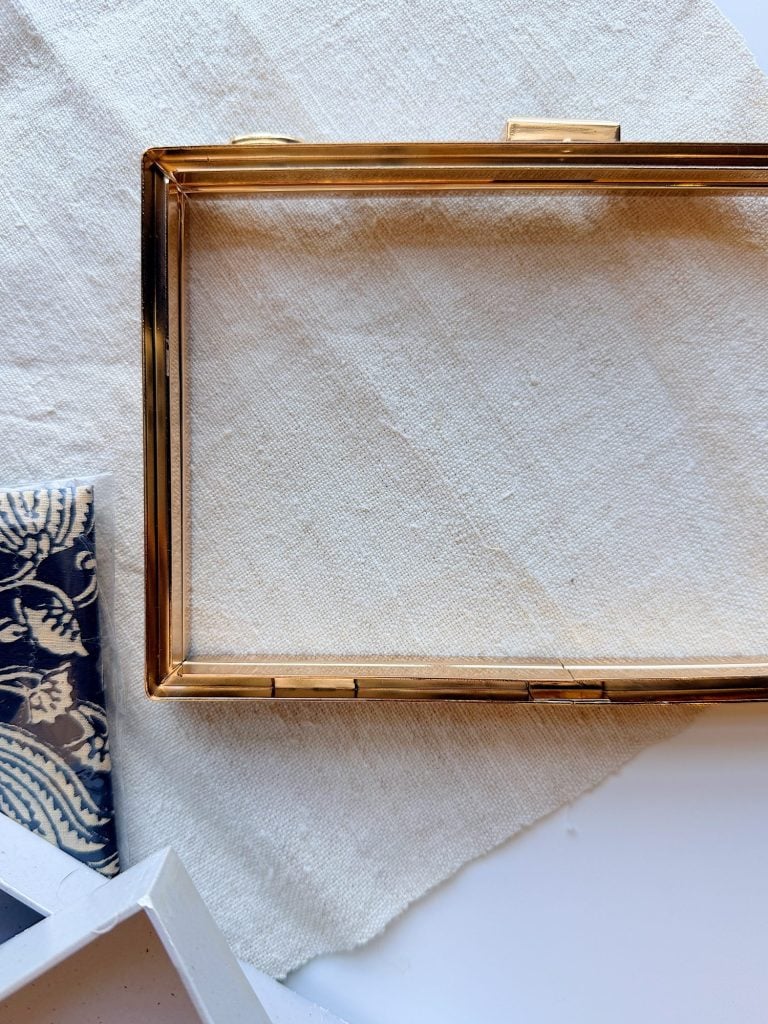 A rectangular gold metal frame rests on a textured beige fabric, with a blue patterned cloth and a white geometric object nearby.