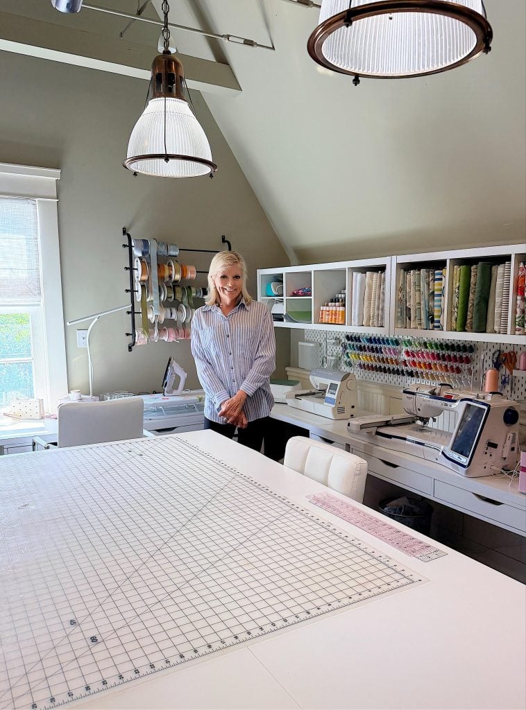 A woman stands in a well-organized sewing room with fabric, ribbons, sewing machines, and tools neatly arranged on shelves and a large cutting table in the foreground.